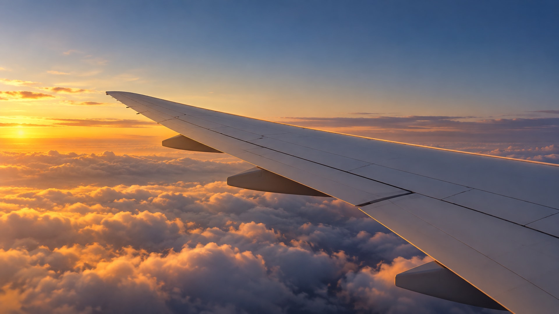Wing of a plain, unmarked airliner above clouds lit orange by the setting sun, seen from a passenger window
